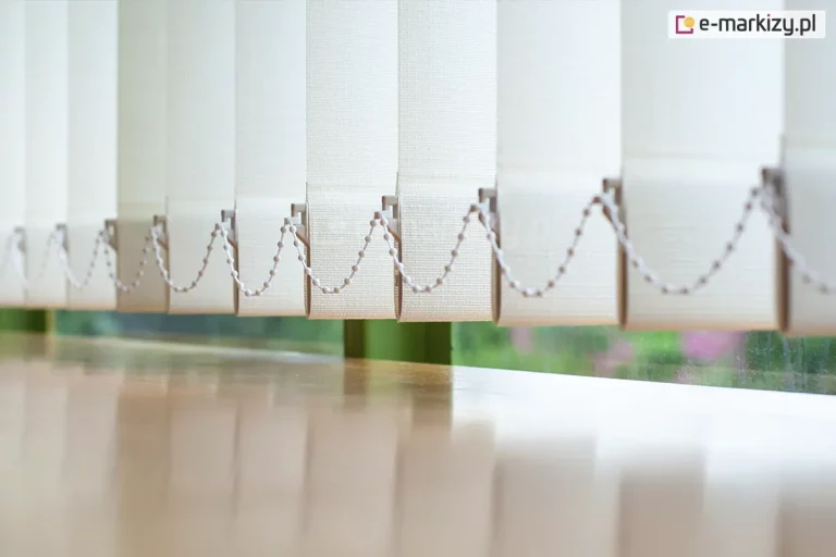 Zbliżenie na dolną część żaluzji pionowych, prezentujące mechanizm stabilizujący lamele nad powierzchnią jasnego, drewnianego blatu. Poszczególne pasy materiału w kolorze kremowym lub bardzo jasnego beżu mają wyraźną strukturę gęstego splotu tkaniny. W dolnej krawędzi każdej lameli znajduje się biały obciążnik, do którego wpięty jest plastikowy łańcuszek koralikowy. Łańcuszek ten łączy wszystkie elementy żaluzji pionowej, zapewniając ich równomierne ułożenie i zapobiegając nadmiernemu kołysaniu. Żaluzje pionowe są ustawione pod kątem, co tworzy rytmiczny, ząbkowany wzór na tle okna. Przez szybę widoczne jest rozmyte, zielone tło roślinności ogrodowej, co sugeruje montaż przesłony w pomieszczeniu z widokiem na naturę. Powierzchnia blatu pod żaluzjami jest gładka i lekko błyszcząca, odbijając pionowe pasy materiału oraz wpadające do wnętrza miękkie światło dzienne.