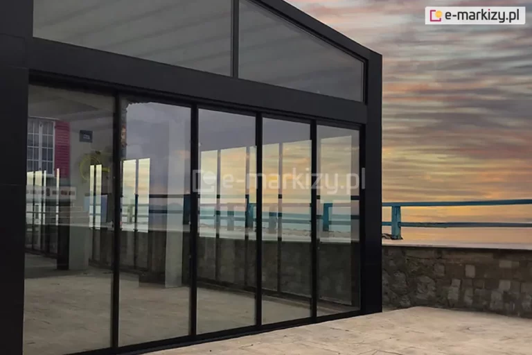 Wielkoformatowa zabudowa patio wykonaną w systemie przesuwnym z czarnymi, eleganckimi profilami aluminiowymi. Centralnym elementem są wysokie, szklane drzwi składające się z pięciu pionowych skrzydeł systemu ramowego Sotro, nad którymi znajduje się stałe przeszklenie w kształcie trapezu, dopasowane do skosu dachu. W taflach szkła odbija się malowniczy widok zachodzącego słońca oraz błękitno-pomarańczowe niebo. Przed zabudową znajduje się taras wyłożony jasnymi płytami kamiennymi, zakończony murkiem z naturalnego kamienia i niebieską balustradą, za którą rozpościera się widok na morze. Wewnątrz przeszklonego pomieszczenia widoczny jest zarys wnętrza z oknem o tradycyjnych szprosach i różowymi okiennicami. Konstrukcja charakteryzuje się minimalistycznym designem i bardzo wąskimi profilami, co maksymalizuje powierzchnię przeszklenia.