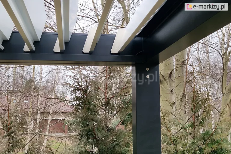 Zbliżenie na wewnętrzny narożnik pergoli lamelowej Sb350 Selt, przedstawiające system osadzenia białych lamel w antracytowej ramie aluminiowej. Zdjęcie prezentuje otwarte pod kątem około 45 stopni aluminiowe lamele dachowe o jasnym wykończeniu, które są zamontowane do bocznej belki nośnej. Widoczna jest precyzyjna szczelina między końcem każdej lameli a ramą, co umożliwia ich swobodny obrót. Konstrukcja wsparta jest na kwadratowym, pionowym słupie w kolorze antracytowym, na którym widoczne są dwa srebrne punkty montażowe (śruby). W samym rogu, przy górnej krawędzi słupa, znajduje się dyskretnie poprowadzony czarny przewód elektryczny, służący prawdopodobnie do zasilania siłownika lub zintegrowanego oświetlenia. Tło ujęcia stanowi naturalna roślinność ogrodowa – pnie brzóz oraz gałęzie krzewów iglastych, a w oddali zarys budynku mieszkalnego. Perspektywa od dołu podkreśla techniczny charakter i wysoką jakość spasowania elementów systemu bioklimatycznego.