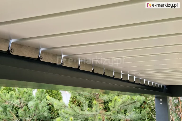 Zbliżenie od dołu na mechanizm obrotowy lamel w nowoczesnej pergoli bioklimatycznej SB350. Zdjęcie przedstawia białe, aluminiowe pióra (lamele) dachu, które są zamontowane do bocznej ramy konstrukcyjnej w kolorze antracytowym. Widoczne są stalowe elementy mocujące oraz system zawiasów, które umożliwiają regulację kąta nachylenia lamel w celu kontroli nasłonecznienia i wentylacji. W dolnej części kadru, pod konstrukcją, widoczne są korony drzew iglastych (sosny) oraz fragment pionowego słupa wsporczego pergoli z widocznymi śrubami montażowymi. Detal ten podkreśla precyzję wykonania ruchomych elementów zadaszenia oraz estetyczne połączenie kolorystyczne bieli lamel z ciemną ramą.