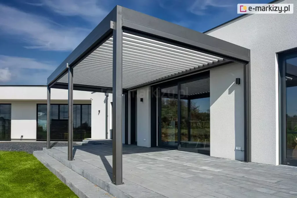 Nowoczesna pergola tarasowa przyścienna SB 400 Aluprof w kolorze antracytowym, zainstalowana na tarasie wyłożonym szarymi płytami betonowymi. Konstrukcja bioklimatyczna Sb400 Selt posiada dach z jasnych, ruchomych lameli w kolorze białym. Pergola przylega do białej, tynkowanej elewacji minimalistycznego budynku o parterowej bryle. W elewacji widoczne są duże przeszklenia w ciemnych ramach, w tym przesuwne drzwi tarasowe. W tle znajduje się fragment drugiego skrzydła domu z oknami osłoniętymi czarnymi żaluzjami fasadowymi. Otoczenie budynku obejmuje zielony trawnik oraz pas ciemnego żwiru wzdłuż fundamentów. Zdjęcie wykonano w pełnym słońcu przy bezchmurnym, niebieskim niebie, co podkreśla czystą formę architektoniczną i kontrastową kolorystykę systemu zadaszenia.