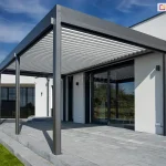 Nowoczesna pergola tarasowa przyścienna SB 400 Aluprof w kolorze antracytowym, zainstalowana na tarasie wyłożonym szarymi płytami betonowymi. Konstrukcja bioklimatyczna Sb400 Selt posiada dach z jasnych, ruchomych lameli w kolorze białym. Pergola przylega do białej, tynkowanej elewacji minimalistycznego budynku o parterowej bryle. W elewacji widoczne są duże przeszklenia w ciemnych ramach, w tym przesuwne drzwi tarasowe. W tle znajduje się fragment drugiego skrzydła domu z oknami osłoniętymi czarnymi żaluzjami fasadowymi. Otoczenie budynku obejmuje zielony trawnik oraz pas ciemnego żwiru wzdłuż fundamentów. Zdjęcie wykonano w pełnym słońcu przy bezchmurnym, niebieskim niebie, co podkreśla czystą formę architektoniczną i kontrastową kolorystykę systemu zadaszenia.