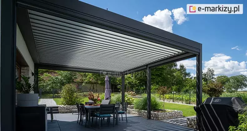 Nowoczesna pergola lamelowa przyścienna Sb400 Aluprof w kolorze antracytowym, zainstalowana na przestronnym tarasie. Zadaszenie składa się z gęsto rozmieszczonych białych lameli, które są ustawione w pozycji lekko uchylonej. Pod pergolą znajduje się strefa jadalniana z dużym stołem i krzesłami z ciemnymi poduszkami. Na krawędzi tarasu widoczny jest profesjonalny grill gazowy osłonięty czarnym pokrowcem. Teren wokół tarasu jest starannie zagospodarowany: widoczne są murki oporowe z gabionów wypełnionych jasnym kamieniem, ozdobne trawy, krzewy oraz zadbany ogród warzywny i drzewa w tle. Zdjęcie wykonano w słoneczny dzień, co podkreśla funkcję ochrony przed słońcem i integrację konstrukcji z naturalnym otoczeniem ogrodu.