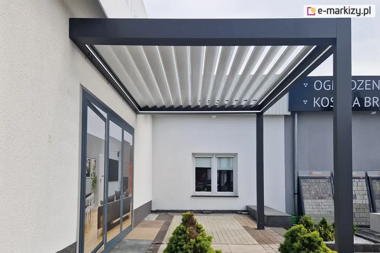 Pergola SB400 Selt przyścienna zamocowana do ściany na ekspozycji sklepu Adams. Antracytowa konstrukcja i białe uchylone lamele tworzą klimatyczne zadaszenie z oświetleniem taśmowym led w górnej opasce konstrukcji. W tle widoczne okno kwadratowe w ścianie i ekspozycja kostki brukowej.