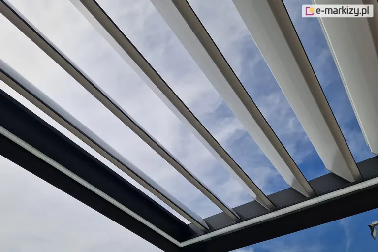 Dach pergoli lamelowej SB400 z białymi lamelami o uchylonej pozycji zamocowanymi do antracytowej konstrukcji z widocznym oświetleniem LED w postaci taśm świetlnych. Przez uchylone lamele widać niebieskie niebo przykryte częściowo chmurami.