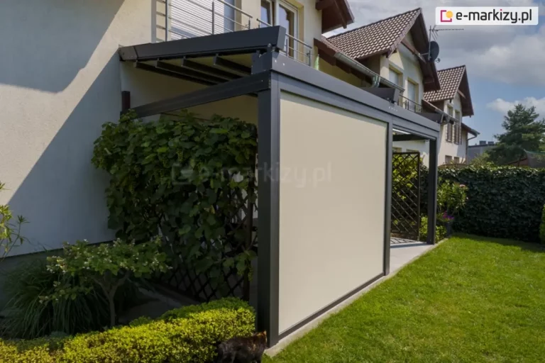 Zdjęcie prezentuje nowoczesną pergolę tarasową model Solid Selt w kolorze antracytowym, zamontowaną do ściany jasnego budynku jednorodzinnego. Głównym elementem wyróżniającym tę realizację jest pionowa przesłona boczna – refleksol przeciwsłoneczny Ziiip Selt w kolorze kremowym, który jest całkowicie opuszczony, wypełniając przestrzeń między słupami konstrukcyjnymi. Zadaszenie pergoli wykonane z segmentowej tkaniny jest częściowo zwinięte w stronę elewacji. Po lewej stronie konstrukcji widoczna jest drewniana kratka porośnięta gęstymi, zielonymi pnączami, a u podstawy znajduje się niski, zadbany żywopłot. Przed pergolą rozciąga się zielony trawnik. W tle widoczne są inne budynki mieszkalne z brązowymi dachami oraz błękitne niebo z lekkimi chmurami. Całość instalacji tworzy osłoniętą i prywatną przestrzeń wypoczynkową na tarasie.