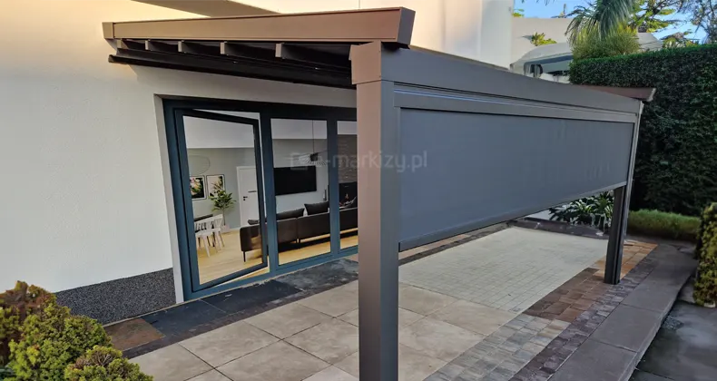 Pergola Square ustawiona przy ścianie o białej elewacji, na tarasie z różnego rodzaju płytek i kostki, wyposażona w refleksol frontowy z antracytową tkaniną.