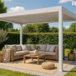 Nowoczesna, wolnostojąca pergola ogrodowa Marlo o wymiarach 4 na 3 metry w kolorze białym, ustawiona na tarasie z jasnoszarych płyt gresowych. Konstrukcja o minimalistycznym designie opiera się na czterech kwadratowych słupach, a jej dach tworzą ruchome lamele aluminiowe, które w tym ujęciu są całkowicie zamknięte. Pod zadaszeniem zaaranżowano przytulną strefę wypoczynkową z dużym narożnikiem z technorattanu w kolorze naturalnego beżu, uzupełnionym o jasne poduszki i pled. W centrum znajduje się niski, prostokątny stolik kawowy oraz pleciony puf. Przestrzeń wypoczynkową wyznacza patio z płyt gresowych, przykryte częściowo plecionym dywanem. Dekorację przestrzeni stanowią rośliny w wiklinowych donicach: drzewko oliwne po lewej stronie oraz strelicja po prawej. Tło aranżacji tworzy gęsty, wysoki żywopłot oraz zadbany trawnik, co podkreśla prywatny i rekreacyjny charakter miejsca.