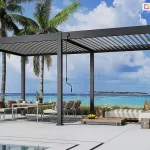 Luksusowa, wolnostojąca pergola tarasowa Marlo, o wymiarach 6 na 3 metry w kolorze antracytowym, zlokalizowana na nadmorskim tarasie. Dwumodułowa konstrukcja opiera się na sześciu słupach, a jej zadaszenie stanowią lamele aluminiowe ustawione pod kątem, co pozwala na częściowy dopływ światła słonecznego. Pod lewym modułem pergoli znajduje się strefa jadalniana z białym stołem i nowoczesnymi krzesłami, natomiast prawy moduł osłania strefę relaksu z beżowym szezlongiem i niskim stolikiem kawowym z owocami. Przy słupach frontowych zamontowane są korby do manualnego sterowania dachem. Taras z jasnoszarych płyt graniczy z niską roślinnością wydmową i szklaną balustradą, za którą rozpościera się widok na turkusowy ocean, białą plażę oraz tropikalną wyspę z palmami na horyzoncie.
