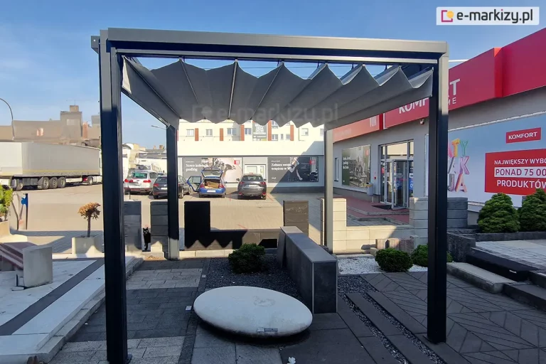Wolnostojąca pergola tarasowa Tako o konstrukcji aluminiowej w kolorze antracytowym, z zadaszeniem z szarej tkaniny rzymskiej, ustawiona na zewnętrznym placu ekspozycyjnym. Materiał dachu jest rozwinięty, tworząc regularne, trapezowe zakładki (fałdy) podwieszone na poprzecznych profilach. Pergola oparta jest na czterech pionowych słupach o kwadratowym przekroju, zamontowanych na podłożu z ciemnych płyt tarasowych i kostki brukowej. Wewnątrz pergoli znajduje się biały, owalny element dekoracyjny przypominający kamień oraz niska roślinność w żwirze. W tle widoczny jest parking z samochodami, duża ciężarówka z naczepą oraz fragmenty budynków handlowych z czerwonymi akcentami i logotypami. Niebo jest bezchmurne, błękitne, a oświetlenie wskazuje na słoneczny dzień.