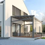 Nowoczesna pergola tarasowa Elegnacy Tarasola o aluminiowej konstrukcji w kolorze ciemnoszarym, przytwierdzona do białej elewacji piętrowego budynku mieszkalnego. Zadaszenie wykonane jest z ruchomej, segmentowej tkaniny w kolorze szarym, a na bocznej ścianie konstrukcji zamontowano pionową osłonę przeciwsłoneczną typu roleta ZIP screen, która jest częściowo opuszczona. Pod pergolą, na podwyższonym tarasie wyłożonym deskami kompozytowymi w kolorze naturalnego drewna, znajduje się strefa jadalniana z dużym stołem i krzesłami ogrodowymi. Budynek posiada nowoczesną architekturę z dużymi oknami tarasowymi oraz zewnętrznymi żaluzjami fasadowymi na piętrze. Teren przed tarasem wybrukowano jasną kostką granitową, a obok widoczny jest fragment zielonego trawnika oraz drzewa i pasmo górskie w oddali pod jasnym, słonecznym niebem.