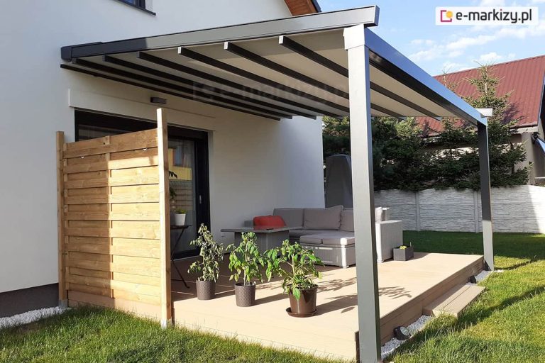Nowoczesna pergola tarasowa o konstrukcji aluminiowej w kolorze antracytowym, zamontowana do białej elewacji piętrowego domu jednorodzinnego. Zadaszenie składa się z beżowej tkaniny rozpiętej na poprzecznych profilach, która jest częściowo zwinięta przy ścianie budynku. Konstrukcja wspiera się na dwóch masywnych, kwadratowych słupach bocznych, a po lewej stronie tarasu zainstalowano pionową ściankę osłonową wykonaną z jasnych, drewnianych desek ułożonych poziomo. Pod pergolą, na podwyższonym tarasie z jasnych desek kompozytowych, znajduje się narożna sofa technorattanowa w kolorze jasnoszarym z pasującymi poduszkami oraz stolik kawowy. Na tarasie ustawiono trzy doniczki z krzewami pomidorów oraz mniejszą skrzynkę z roślinami. Taras graniczy z zielonym trawnikiem, a w tle widoczne są iglaki, biały betonowy płot oraz fragment sąsiedniego budynku z czerwonym dachem. Zdjęcie wykonane w świetle dziennym przy lekko zachmurzonym niebie.