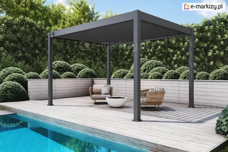 Nowoczesna, wolnostojąca pergola lamelowa w kolorze antracytowym, zainstalowana na tarasie z jasnych desek drewnopodobnych tuż przy krawędzi basenu. Konstrukcja wspiera się na czterech narożnych słupach o kwadratowym przekroju, a jej dach tworzą ruchome lamele aluminiowe ustawione w pozycji zamkniętej. Pod zadaszeniem znajduje się minimalistyczna strefa wypoczynkowa z dwoma fotelami o beżowym splocie, wyposażonymi w jasne poduszki, oraz niskie palenisko ogrodowe w formie białej misy. Taras jest otoczony niskim murkiem z poziomych paneli w jasnym kolorze, za którym rozciąga się gęsty żywopłot z formowanych w kule krzewów liściastych. W lewym dolnym rogu widoczna jest krystalicznie czysta, błękitna woda basenu, w której odbijają się fragmenty architektury i roślinności. Przy jednym ze słupów pergoli zamontowany jest mechanizm korbowy służący do ręcznej regulacji kąta nachylenia lameli dachowych.