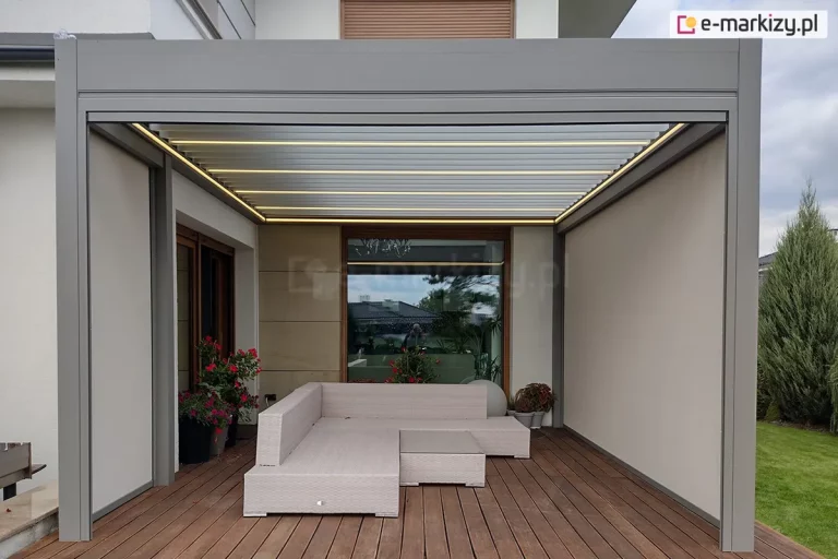 Pergola tarasowa z dachem lamelowym w jasnym kolorze ustawiona nad drewnianym tarasem, na którym ustawiono ratanowe meble ogrodowe oraz rośliny w doniczkach. Dwie ściany prawa i lewa osłonięte zostały przez refleksol ZIP L ZIP U, który skutecznie chroni taras przed słońcem, deszczem i śniegiem oraz wiatrem. W tle tarasu duże okno, w którym odbija się taras oraz dom sąsiada. Z prawej strony za tarasem widoczna roślina zielona z iglastymi gałązkami oraz trawnik.