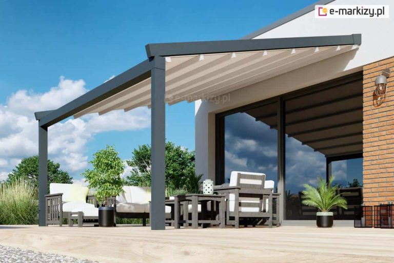 Nowoczesna przyścienna pergola tarasowa o konstrukcji aluminiowej w kolorze antracytowym, zamontowana do białej elewacji budynku z elementami dekoracyjnej cegły. Zadaszenie stanowi jasnoszary, ruchomy dach tkaninowy, który jest całkowicie rozłożony na profilach poprzecznych. Pod pergolą, na drewnianym tarasie z jasnych desek, znajduje się zestaw mebli ogrodowych składający się z foteli i stolików o grafitowej konstrukcji z białymi poduszkami. Duże, przeszkolone drzwi tarasowe odbijają otoczenie, a cała strefa wypoczynku jest udekorowana roślinami w donicach, w tym małą palmą. W tle widoczne jest bezchmurne, błękitne niebo oraz niska roślinność ogrodowa, co podkreśla minimalistyczny i elegancki design zadaszenia.
