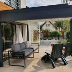 Zdjęcie prezentuje widok spod nowoczesnej pergoli aluminiowej z dachem tkaninowym Luxury w kolorze antracytowym, która została zamontowana jako przedłużenie strefy wypoczynkowej przy nowoczesnym budynku. Konstrukcja posiada płaski dach z białej wodoodpornej tkaniny rozciągniętej pomiędzy białymi belkami przeciwwietrznymi. Pod zadaszeniem, na jasnych płytkach tarasowych, ustawiono komfortową, ciemnoszarą sofę ogrodową z miękkimi poduszkami oraz dwa fotele rozkładane z ciemnozielonego tworzywa. Pergola przylega do jasnej elewacji domu, wzbogaconej o detale z naturalnego drewna i duże przeszklenia tarasowe. W tle widoczna jest starannie zaprojektowana roślinność ogrodowa, w tym drzewka formowane na pniu oraz biała pergola kratkowa z pnącymi kwiatami, a dalej fragmenty sąsiedniej zabudowy jednorodzinnej. Solidne profile aluminiowe tworzą wyraźną ramę widokową na ogród, podkreślając funkcjonalność i estetykę nowoczesnej zabudowy tarasu.