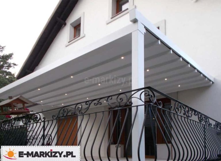 Biała przyścienna pergola tarasowa zainstalowana nad balkonem z ozdobną, kutą balustradą o falistym wzorze. Konstrukcja posiada zadaszenie z jasnej tkaniny rzymskiej ułożonej w charakterystyczne fale, w którą wbudowano punktowe oświetlenie LED rozmieszczone regularnie na profilach poprzecznych. Pergola wspiera się na solidnym, białym słupie o kwadratowym przekroju i jest zamontowana bezpośrednio do elewacji piętrowego domu z widocznymi oknami w drewnianych ramie. W lewym dolnym rogu, przy balustradzie, widoczny jest fragment drewnianego karmnika dla ptaków. Zdjęcie wykonane z perspektywy dołu, prezentuje estetyczne wykończenie systemu zadaszenia oraz funkcjonalne wykorzystanie oświetlenia zewnętrznego, które pozwala na korzystanie z tarasu po zmroku.