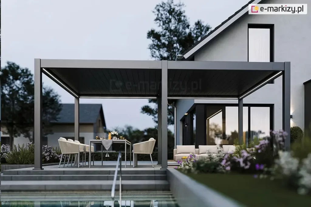 Wolnostojąca pergola bioklimatyczna Rubio dwumodułowa ustawiona na sześciu solidnych podporach przy ścianie budynku. Pergola w kolorze antracytowym, stanowiąca przedłużenie strefy wypoczynkowej nowoczesnego domu jednorodzinnego. Aluminiowa konstrukcja z zamkniętym dachem lamelowym wyposażona jest w liniowe oświetlenie LED, które rzuca jasne światło na przestrzeń pod zadaszeniem. Pod pergolą znajduje się strefa jadalniana z szarym stołem i jasnymi fotelami oraz strefa lounge z sofą. Całość umieszczona na podwyższonym, betonowym tarasie, do którego prowadzą szerokie stopnie. W bezpośrednim sąsiedztwie widoczny jest basen z metalową poręczą oraz starannie zaprojektowany ogród: kwitnące rabaty z fioletowymi kwiatami, niska roślinność ozdobna oraz drzewa liściaste w tle pod zachmurzonym niebem o zmierzchu. Bryła domu z białym tynkiem i ciemnymi ramami okiennymi tworzy spójną całość z kolorystyką pergoli, a zapalone oświetlenie wewnętrzne i zewnętrzne budynku podkreśla luksusowy charakter realizacji.