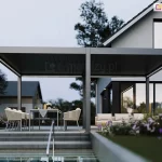 Wolnostojąca pergola bioklimatyczna Rubio dwumodułowa ustawiona na sześciu solidnych podporach przy ścianie budynku. Pergola w kolorze antracytowym, stanowiąca przedłużenie strefy wypoczynkowej nowoczesnego domu jednorodzinnego. Aluminiowa konstrukcja z zamkniętym dachem lamelowym wyposażona jest w liniowe oświetlenie LED, które rzuca jasne światło na przestrzeń pod zadaszeniem. Pod pergolą znajduje się strefa jadalniana z szarym stołem i jasnymi fotelami oraz strefa lounge z sofą. Całość umieszczona na podwyższonym, betonowym tarasie, do którego prowadzą szerokie stopnie. W bezpośrednim sąsiedztwie widoczny jest basen z metalową poręczą oraz starannie zaprojektowany ogród: kwitnące rabaty z fioletowymi kwiatami, niska roślinność ozdobna oraz drzewa liściaste w tle pod zachmurzonym niebem o zmierzchu. Bryła domu z białym tynkiem i ciemnymi ramami okiennymi tworzy spójną całość z kolorystyką pergoli, a zapalone oświetlenie wewnętrzne i zewnętrzne budynku podkreśla luksusowy charakter realizacji.