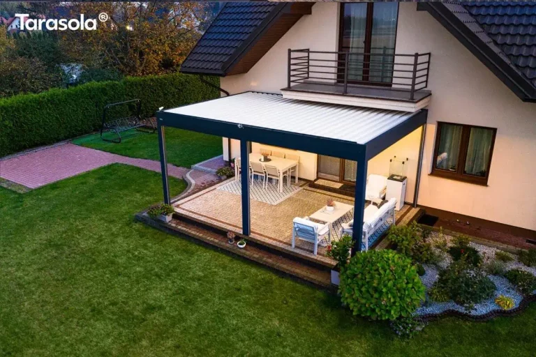 Zdjęcie wykonane z góry przedstawia nowoczesną, dwumodułową pergolę Essential Tarasola zainstalowaną przy elewacji domu jednorodzinnego. Konstrukcja modelu Essential Tarasola wykonana jest z profili aluminiowych w kolorze antracytowym, a jej zadaszenie stanowią białe lamele, które w tej perspektywie tworzą jednolitą, jasną powierzchnię dachu. Pergola jest zamontowana pod balkonem z ciemnobrązową balustradą i przylega do jasnej, beżowej ściany budynku. Taras pod pergolą Essential Tarasola został wyłożony jasną kostką brukową z dekoracyjnym, ciemniejszym obramowaniem, a cała strefa wypoczynkowa jest oświetlona ciepłym, zintegrowanym światłem, które wydobywa się spod dachu konstrukcji. Wewnątrz lewego segmentu znajduje się jadalnia z prostokątnym białym stołem i krzesłami ustawionymi na jasnym dywanie. Prawy segment pełni funkcję salonową, wyposażoną w sofę ogrodową, dwa fotele oraz stolik kawowy, również umieszczone na dywanie o geometrycznym wzorze. Po prawej stronie tarasu, obok mebli, widoczny jest pionowy, biały ogrzewacz tarasowy. Całość konstrukcji Essential Tarasola otoczona jest zadbanym ogrodem z zielonym trawnikiem, po prawej stronie znajduje się ozdobna rabata z krzewami i hortensjami wysypana szarym gresem, a w tle widać ścieżkę z czerwonej kostki prowadzącą w stronę huśtawki ogrodowej. Budynek posiada skośny dach pokryty ciemną dachówką oraz brązową stolarkę okienną.