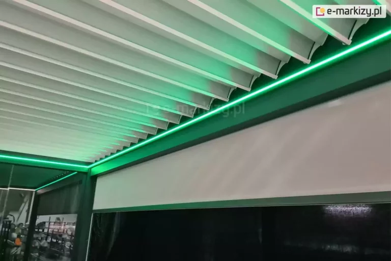 Zdjęcie prezentuje szczegółowe ujęcie wewnętrznej konstrukcji nowoczesnej pergoli tarasowej z dachem lamelowym, wykonane z perspektywy dolnej. Głównym elementem zadaszenia są białe, aluminiowe lamele ułożone równolegle obok siebie, które na krawędziach posiadają charakterystyczne wyprofilowanie ułatwiające odprowadzanie wody. Wzdłuż wewnętrznej ramy konstrukcyjnej, na styku z dachem, zamontowano liniowe oświetlenie LED, które emituje intensywne światło o nasyconej, zielonej barwie. Zielona poświata oświetla krawędzie lameli oraz górną część bocznej osłony. Poniżej linii światła widoczna jest pionowa, jasna roleta typu screen, która jest częściowo rozwinięta, a jej powierzchnia delikatnie odbija zielony blask. Rama pergoli ma ciemny, prawdopodobnie antracytowy kolor, który kontrastuje z jasnymi lamelami i jaskrawym oświetleniem. W lewym dolnym rogu zdjęcia, przez przeszklony fragment konstrukcji, prześwituje zarysy wnętrza pomieszczenia lub hali wystawowej, sugerując, że zdjęcie mogło zostać wykonane w salonie ekspozycyjnym.