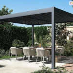 Nowoczesna wolnostojąca pergola ogrodowa o aluminiowej konstrukcji w kolorze antracytowym, usytuowana na tarasie z jasnych płyt betonowych. Zadaszenie pergoli Marlo Duo wykonane jest z poziomych, ruchomych lameli, które są lekko uchylone, tworząc charakterystyczny liniowy cień na posadzce tarasu. Konstrukcja opiera się na czterech pionowych, kwadratowych słupach. Pod pergolą znajduje się strefa jadalniana z dużym stołem o drewnianym blacie, przy którym ustawiono eleganckie fotele ogrodowe. Fotele posiadają ażurowe oparcia wykonane z jasnego, plecionego sznurka oraz szare poduszki siedziskowe. Na stole widoczna jest dekoracyjna zastawa stołowa. Tło stanowi gęsty, wysoki żywopłot oraz korony drzew pod bezchmurnym, błękitnym niebem. Po prawej stronie, na ziemi, widoczny jest wyraźny pas cienia rzucanego przez konstrukcję zadaszenia, który pada na fragment nawierzchni z czerwonej kostki brukowej. Całość aranżacji podkreśla nowoczesny design i funkcjonalność przydomowej przestrzeni rekreacyjnej.