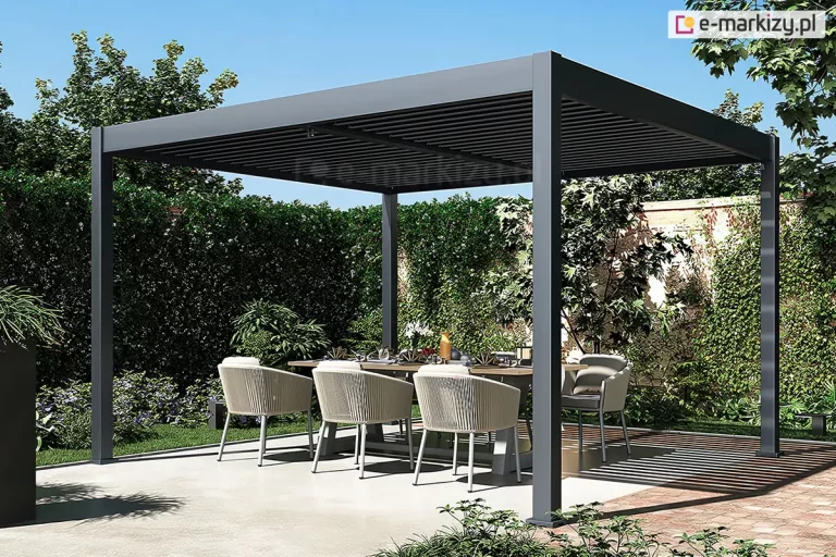 Nowoczesna wolnostojąca pergola ogrodowa o aluminiowej konstrukcji w kolorze antracytowym, usytuowana na tarasie z jasnych płyt betonowych. Zadaszenie pergoli Marlo Duo wykonane jest z poziomych, ruchomych lameli, które są lekko uchylone, tworząc charakterystyczny liniowy cień na posadzce tarasu. Konstrukcja opiera się na czterech pionowych, kwadratowych słupach. Pod pergolą znajduje się strefa jadalniana z dużym stołem o drewnianym blacie, przy którym ustawiono eleganckie fotele ogrodowe. Fotele posiadają ażurowe oparcia wykonane z jasnego, plecionego sznurka oraz szare poduszki siedziskowe. Na stole widoczna jest dekoracyjna zastawa stołowa. Tło stanowi gęsty, wysoki żywopłot oraz korony drzew pod bezchmurnym, błękitnym niebem. Po prawej stronie, na ziemi, widoczny jest wyraźny pas cienia rzucanego przez konstrukcję zadaszenia, który pada na fragment nawierzchni z czerwonej kostki brukowej. Całość aranżacji podkreśla nowoczesny design i funkcjonalność przydomowej przestrzeni rekreacyjnej.