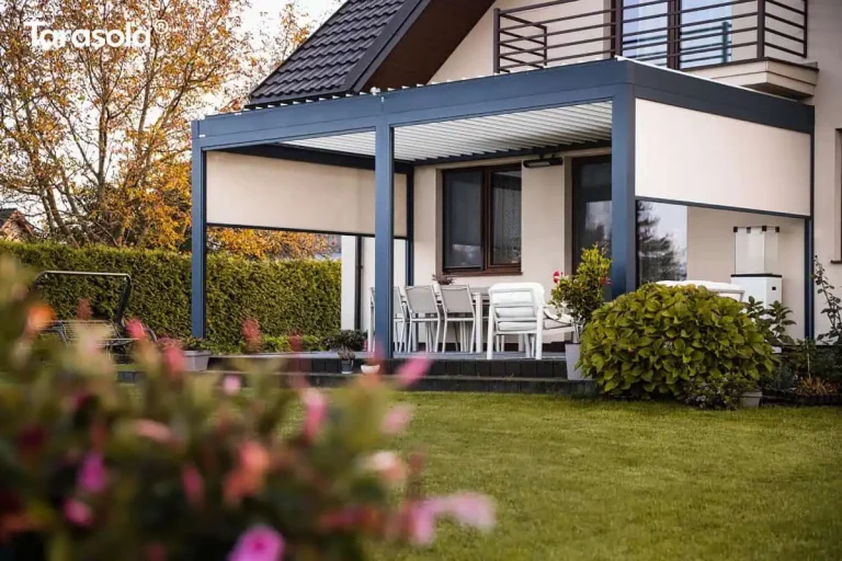 Perspektywa ogrodowa przedstawiająca nowoczesną pergolę Essential Tarasola o konstrukcji w kolorze antracytowym, zamontowaną przy elewacji jasnego domu jednorodzinnego. Zadaszenie Essential Tarasola posiada dach wykonany z białych lameli, które są w tym momencie zamknięte, tworząc szczelną i płaską powierzchnię. Konstrukcja opiera się na solidnych profilach aluminiowych, w tym na centralnym słupie dzielącym taras na dwie funkcjonalne sekcje. W obu segmentach zamontowano pionowe rolety zewnętrzne typu screen w kolorze beżowym; lewa roleta jest opuszczona do połowy wysokości, natomiast prawa jest niemal całkowicie podniesiona, co obrazuje zakres regulacji osłon. Pod pergolą Essential Tarasola znajduje się strefa jadalniana z jasnym stołem i krzesłami oraz część wypoczynkowa z białymi fotelami ogrodowymi. Na ścianie budynku, pod dachem konstrukcji, widoczny jest zamontowany promiennik ciepła, a po prawej stronie tarasu stoi biały ogrzewacz gazowy z szklaną obudową płomienia. Cały taras jest podwyższony względem ogrodu, wyłożony szarą kostką i wykończony ciemnym obrzeżem. Na pierwszym planie, w dolnym lewym rogu, widoczne są nieostre, różowe kwiaty, które nadają zdjęciu głębi. W tle po lewej stronie znajduje się wysoki, zielony żywopłot oraz huśtawka ogrodowa na trawniku. Dom posiada jasną elewację, brązową stolarkę okienną oraz ciemny, skośny dach i balkon z balustradą znajdujący się powyżej linii pergoli.