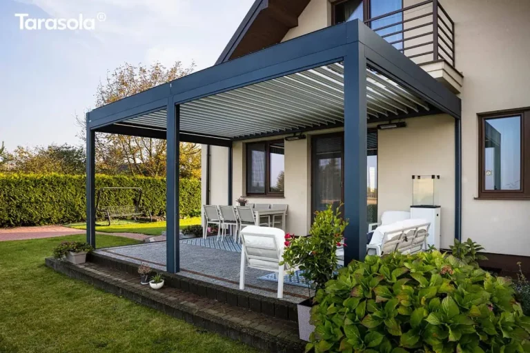 Zdjęcie przedstawia szeroką perspektywę nowoczesnego zadaszenia tarasowego Essential Tarasola zamontowanego do elewacji piętrowego domu jednorodzinnego. Konstrukcja w kolorze ciemnoantracytowym opiera się na trzech masywnych słupach, które dzielą przestrzeń tarasu na dwa segmenty. Dach pergoli Essential Tarasola tworzą białe, aluminiowe lamele, które są częściowo uchylone, co pozwala na regulację dopływu światła i naturalną wentylację przestrzeni pod zadaszeniem. Taras jest wyniesiony ponad poziom trawnika i wykończony szarą kostką brukową oraz ciemną palisadą betonową na krawędziach. Pod pergolą Essential Tarasola wydzielono dwie strefy funkcjonalne: jadalnianą z dużym białym stołem i sześcioma krzesłami oraz relaksacyjną z białymi, niskimi fotelami ogrodowymi ustawionymi na geometrycznych dywanach zewnętrznych. Na jasnej ścianie budynku, pod dachem konstrukcji, widoczne są dwa promienniki ciepła, które umożliwiają komfortowe korzystanie z tarasu w chłodniejsze wieczory. Po lewej stronie, w tle ogrodu, znajduje się klasyczna huśtawka ogrodowa oraz wysoki, gęsty żywopłot oddzielający posesję. Na pierwszym planie po prawej stronie widoczne są duże, zielone krzewy hortensji w donicach. Budynek posiada jasną, kremową elewację, brązową stolarkę okienną oraz balkon z metalową balustradą znajdujący się bezpośrednio nad częścią konstrukcji Essential Tarasola.