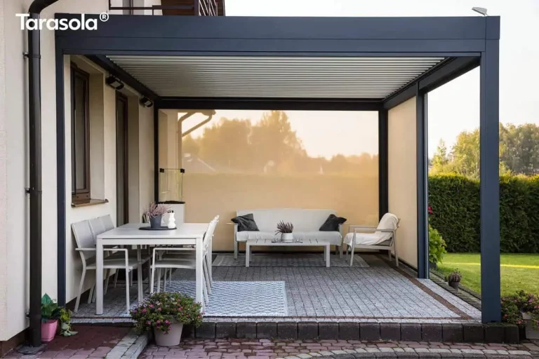 Nowoczesna aluminiowa pergola tarasowa Essential Tarasola w eleganckim, ciemnoantracytowym kolorze, zamontowana jako konstrukcja przyścienna do elewacji budynku. Model Essential Tarasola charakteryzuje się płaskim dachem z ruchomymi lamelami, które na zdjęciu są ustawione w pozycji zamkniętej, tworząc szczelną barierę chroniącą przed słońcem i deszczem. Konstrukcja wspiera się na solidnych, kwadratowych słupach bocznych, które precyzyjnie wyznaczają strefę wypoczynku. Lewa strona oraz tył pergoli zostały wyposażone w jasne przesłony przeciwsłoneczne typu screen, które przepuszczają miękkie, rozproszone światło, jednocześnie odgradzając wnętrze tarasu od otoczenia i wiatru. Pod zadaszeniem Essential Tarasola znajduje się starannie zaaranżowana przestrzeń podzielona na część jadalnianą i salonową, ułożona na szarej kostce brukowej z ozdobnym obramowaniem z ciemniejszej kostki. Po lewej stronie ustawiono biały stół ogrodowy z czterema dopasowanymi kolorystycznie krzesłami o lekkiej konstrukcji, umieszczony na niebiesko-białym dywanie zewnętrznym w geometryczne wzory. W głębi tarasu, pod tylną przesłoną screen, znajduje się strefa relaksu z białą sofą ogrodową udekorowaną ciemnymi poduszkami, niskim stolikiem kawowym oraz pojedynczym fotelem z jasnym narzutem, również ustawionymi na ozdobnym dywanie. Na jasnej elewacji domu widoczne są brązowe ramy okienne, czarna rura spustowa oraz zamontowane pod dachem pergoli oświetlenie zewnętrzne w formie punktowych reflektorów. Na pierwszym planie, przy krawędzi tarasu wykończonego ciemną palisadą, stoi donica z kwitnącymi różowymi kwiatami, a po prawej stronie rozciąga się zielony ogród z przystrzyżonym żywopłotem i trawnikiem. Całość aranżacji podkreśla nowoczesny design produktu Essential Tarasola, łącząc funkcjonalność z minimalistyczną estetyką.