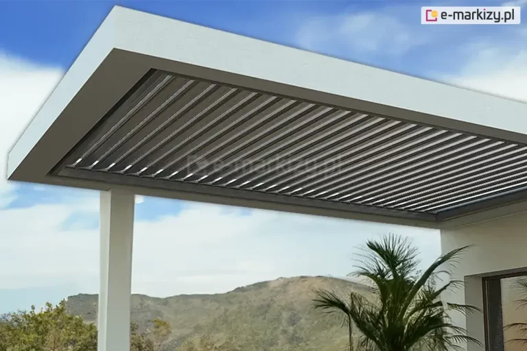 Zdjęcie prezentuje nowoczesną pergolę dachową SB400R w widoku od dołu, zamontowaną jako przedłużenie zadaszenia budynku. Konstrukcja posiada aluminiowe lamele w kolorze antracytowym, które są ustawione w pozycji lekko uchylonej, co pozwala na prześwity światła dziennego i naturalną cyrkulację powietrza. Rama zadaszenia oraz podtrzymujący ją słup nośny są wykonane w kolorze białym, co tworzy kontrastowy, minimalistyczny wygląd. Pergola jest zintegrowana z bryłą nowoczesnego domu, z widocznym fragmentem białej elewacji oraz przeszklonymi drzwiami. W tle rozpościera się panoramiczny widok na zielone wzgórza pod jasnobłękitnym niebem z rozproszonymi chmurami. Na tarasie, w pobliżu konstrukcji, widoczna jest ozdobna palma doniczkowa, podkreślająca rekreacyjny charakter miejsca.