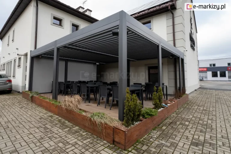 Dwumodułowa pergola tarasowa SB400 w kolorze antracytowym, zamontowana jako zadaszenie zewnętrzne przy jasnej elewacji piętrowego budynku. Konstrukcja bioklimatyczna posiada dach z uchylnych lameli ustawionych w pozycji zamkniętej. Od strony bocznej zainstalowano pełną przesłonę pionową (screen) w kolorze szarym, zapewniającą osłonę przed wiatrem i wzrokiem osób postronnych. Pod pergolą, na drewnianym podeście, znajduje się strefa gastronomiczna z czarnymi, prostokątnymi stołami i dopasowanymi krzesłami. Całość została odseparowana od parkingu z kostki brukowej za pomocą wysokiej, narożnej donicy wykonanej z brązowych desek, w której posadzono ozdobne krzewy iglaste. W tle widoczny jest fragment srebrnego samochodu po lewej, a z prawej widoczna elewacja sąsiedniego budynku.