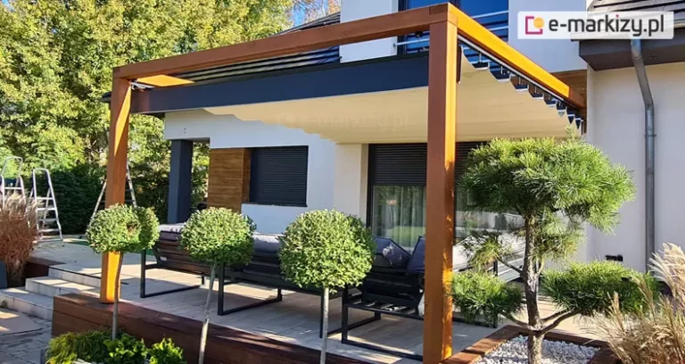 Pergola drewniana koro wolnostojąca z dachem supro z napędem elektrycznym wpasowana w elegancką, jasną fasadę o drewnianych elementach dekoracyjnych. Pod dachem pergoli ustawiono meble tarasowe z metalową ramą oraz szarymi poduchami. Wokół tarasu posadzono liczne drzewka. W tle widoczne drzewa liściaste.