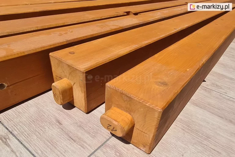 Ułożone na płytkach gresowych o jasnym drewnianym wzorze belki drewniane do zbudowania konstrukcji pergoli. Na dwóch centralnie ułożonych belkach widoczne czopy do łączenia elementów.