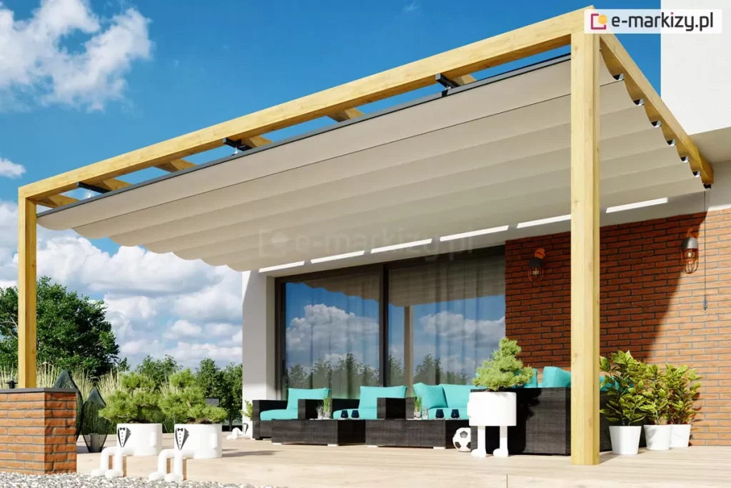 Pergola drewniana Juko przyścienna o konstrukcji nośnej wykonanej z jasnego drewna, zamontowana bezpośrednio do ściany budynku wykończonej czerwoną cegłą. Pergola posiada zadaszenie w formie rozsuwanego materiału w kolorze kremowym, który jest ułożony w równoległe fałdy zamocowane na prowadnicach. Konstrukcja wsparta jest na dwóch pionowych drewnianych słupach ustawionych na krawędzi tarasu, wyłożonego jasnymi płytami nawierzchniowymi. Pod zadaszeniem znajduje się zestaw wypoczynkowy składający się z ciemnych mebli z jasnoniebieskimi poduszkami, a w przestrzeni przed pergolą ustawione są liczne donice z zielonymi roślinami ozdobnymi. Tło zdjęcia stanowi fragment nowoczesnego domu z dużym przeszkleniem oraz czyste, błękitne niebo z delikatnymi chmurami.