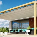 Pergola drewniana Juko przyścienna o konstrukcji nośnej wykonanej z jasnego drewna, zamontowana bezpośrednio do ściany budynku wykończonej czerwoną cegłą. Pergola posiada zadaszenie w formie rozsuwanego materiału w kolorze kremowym, który jest ułożony w równoległe fałdy zamocowane na prowadnicach. Konstrukcja wsparta jest na dwóch pionowych drewnianych słupach ustawionych na krawędzi tarasu, wyłożonego jasnymi płytami nawierzchniowymi. Pod zadaszeniem znajduje się zestaw wypoczynkowy składający się z ciemnych mebli z jasnoniebieskimi poduszkami, a w przestrzeni przed pergolą ustawione są liczne donice z zielonymi roślinami ozdobnymi. Tło zdjęcia stanowi fragment nowoczesnego domu z dużym przeszkleniem oraz czyste, błękitne niebo z delikatnymi chmurami.
