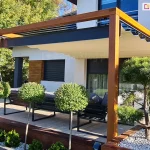 Pergola drewniana koro przyścienna z dachem supro z napędem elektrycznym wpasowana w elegancką, jasną fasadę o drewnianych elementach dekoracyjnych. Pod dachem ustawiono meble wypoczynkowe, wokół tarasu ze schodkami prowadzącymi do ogrodu posadzono kilka drzewek.