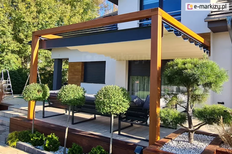 Pergola drewniana koro przyścienna z dachem supro z napędem elektrycznym wpasowana w elegancką, jasną fasadę o drewnianych elementach dekoracyjnych. Pod dachem ustawiono meble wypoczynkowe, wokół tarasu ze schodkami prowadzącymi do ogrodu posadzono kilka drzewek.