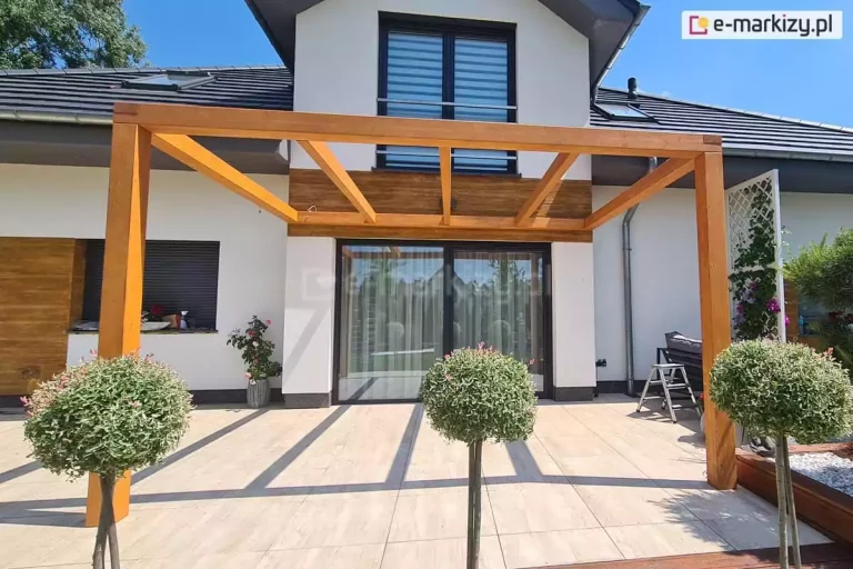 Pergola drewniana Juko przyścienna pokryta lakierobejcą z trzema wewnętrznymi krokwiami ustawiona na tarasie przy domu o białej elewacji z dużym oknem tarasowym o antracytowej ramie. Na pierwszym planie trzy niskie drzewka o kulisto przystrzyżonych koronach na cienkich pniach.