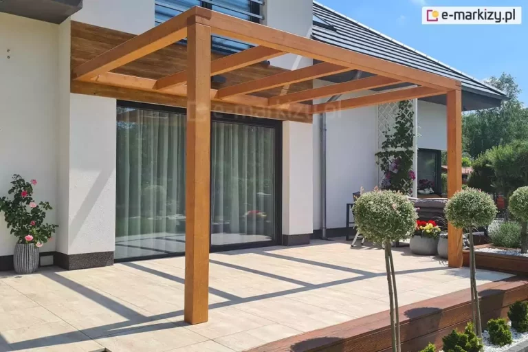 Pergola drewniana przyścienna Juko stanowiąca częściowe zadaszenie tarasu z jasnej płytki gresowej, na którym ustawiono wyłącznie doniczkę z kwiatami. W otoczeniu tarasu widoczne liczne rośliny.