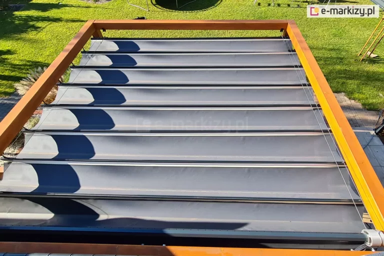 Pergola drewniana Koro z rozwiniętym dachem Supro z tkaniną Opatex o antracytowym kolorze od zewnątrz na tarasie z widocznym zielonym trawnikiem.