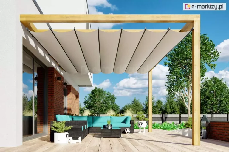 Pergola drewniana Juko nad drewnianym tarasem z meblami w morskim kolorze i donicami z kwiatami. W tle widoczna brzoza z zielonymi liśćmi oraz inne drzewa, a także lekko przykryte białymi chmurami niebieskie niebo.