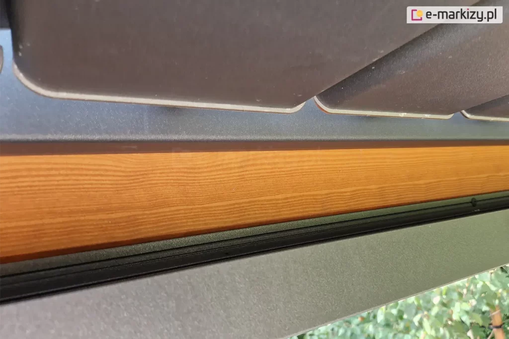 techniczne zbliżenie na system odprowadzania wody w pergoli z dachem lamelowym Selt SB400. Widoczny jest detal montażowy, w którym czarna, aluminiowa rynna została umiejscowiona bezpośrednio pod krawędzią antracytowych lameli dachu. Rynna jest zamontowana do bocznej płaszczyzny solidnej, drewnianej belki konstrukcyjnej o wyraźnym rysunku słojów i miodowym odcieniu. Lamele dachu są lekko uchylone, co pokazuje ich profil oraz sposób, w jaki woda opadowa jest kierowana z ich powierzchni do systemu rynnowego. Konstrukcja zapewnia szczelność i ochronę drewnianych elementów przed bezpośrednim kontaktem z wilgocią. W tle, pod dolną krawędzią rynny, widoczne są rozmyte zielone liście drzew ogrodowych.