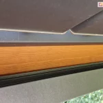 techniczne zbliżenie na system odprowadzania wody w pergoli z dachem lamelowym Selt SB400. Widoczny jest detal montażowy, w którym czarna, aluminiowa rynna została umiejscowiona bezpośrednio pod krawędzią antracytowych lameli dachu. Rynna jest zamontowana do bocznej płaszczyzny solidnej, drewnianej belki konstrukcyjnej o wyraźnym rysunku słojów i miodowym odcieniu. Lamele dachu są lekko uchylone, co pokazuje ich profil oraz sposób, w jaki woda opadowa jest kierowana z ich powierzchni do systemu rynnowego. Konstrukcja zapewnia szczelność i ochronę drewnianych elementów przed bezpośrednim kontaktem z wilgocią. W tle, pod dolną krawędzią rynny, widoczne są rozmyte zielone liście drzew ogrodowych.