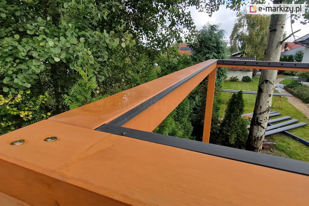 zbliżenie na górną część konstrukcji pergoli drewnianej podczas montażu systemu dachu lamelowego SB400MD. Na pierwszym planie widoczny jest narożnik solidnej ramy wykonanej z drewna klejonego w ciepłym, miodowym odcieniu, na której zamontowano czarne, aluminiowe listwy obwodowe. Listwy te są precyzyjnie połączone pod kątem prostym i przykręcone do drewna, tworząc bazę pod przyszły montaż lameli. Na powierzchni drewna widoczne są śruby montażowe oraz krople deszczu, co świadczy o odporności materiałów na warunki atmosferyczne. W tle rozpościera się widok na ogród z gęstą roślinnością, drzewami liściastymi oraz fragmentem trawnika, na którym leżą przygotowane do montażu profile aluminiowe. Perspektywa z góry pozwala na dokładne przyjrzenie się detalom wykończenia i integracji elementów metalowych z drewnianą konstrukcją nośną.