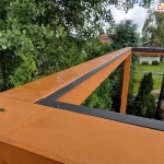 zbliżenie na górną część konstrukcji pergoli drewnianej podczas montażu systemu dachu lamelowego SB400MD. Na pierwszym planie widoczny jest narożnik solidnej ramy wykonanej z drewna klejonego w ciepłym, miodowym odcieniu, na której zamontowano czarne, aluminiowe listwy obwodowe. Listwy te są precyzyjnie połączone pod kątem prostym i przykręcone do drewna, tworząc bazę pod przyszły montaż lameli. Na powierzchni drewna widoczne są śruby montażowe oraz krople deszczu, co świadczy o odporności materiałów na warunki atmosferyczne. W tle rozpościera się widok na ogród z gęstą roślinnością, drzewami liściastymi oraz fragmentem trawnika, na którym leżą przygotowane do montażu profile aluminiowe. Perspektywa z góry pozwala na dokładne przyjrzenie się detalom wykończenia i integracji elementów metalowych z drewnianą konstrukcją nośną.