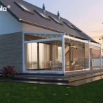 Nowoczesna, biała pergola tarasowa Elegancy Tarasola o konstrukcji aluminiowej z rozsuwanym dachem tkaninowym oraz pełną zabudową szklaną, zamontowana przy elewacji współczesnego domu jednorodzinnego. Przeszklone ściany boczne w systemie przesuwnym tworzą zamknięty ogród letni, wewnątrz którego zaaranżowano strefę wypoczynkową z sofami, fotelami i drewnianym stolikiem kawowym. W profilach dachowych zintegrowano punktowe oświetlenie LED, a na belce konstrukcyjnej zamontowano elektryczny promiennik ciepła, co umożliwia korzystanie z tarasu po zmroku i w chłodniejsze dni. Budynek posiada elewację wykończoną jasnoszarymi panelami poziomymi oraz wysoki, spadzisty dach pokryty ciemną dachówką z oknami połaciowymi i kominkami wentylacyjnymi. Całość uwieczniona w scenerii zachodzącego słońca, na tle zielonego trawnika, krzewów róż oraz szerokich, ciemnych stopni tarasowych prowadzących do ogrodu.