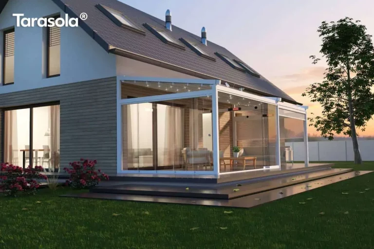 Nowoczesna, biała pergola tarasowa Elegancy Tarasola o konstrukcji aluminiowej z rozsuwanym dachem tkaninowym oraz pełną zabudową szklaną, zamontowana przy elewacji współczesnego domu jednorodzinnego. Przeszklone ściany boczne w systemie przesuwnym tworzą zamknięty ogród letni, wewnątrz którego zaaranżowano strefę wypoczynkową z sofami, fotelami i drewnianym stolikiem kawowym. W profilach dachowych zintegrowano punktowe oświetlenie LED, a na belce konstrukcyjnej zamontowano elektryczny promiennik ciepła, co umożliwia korzystanie z tarasu po zmroku i w chłodniejsze dni. Budynek posiada elewację wykończoną jasnoszarymi panelami poziomymi oraz wysoki, spadzisty dach pokryty ciemną dachówką z oknami połaciowymi i kominkami wentylacyjnymi. Całość uwieczniona w scenerii zachodzącego słońca, na tle zielonego trawnika, krzewów róż oraz szerokich, ciemnych stopni tarasowych prowadzących do ogrodu.