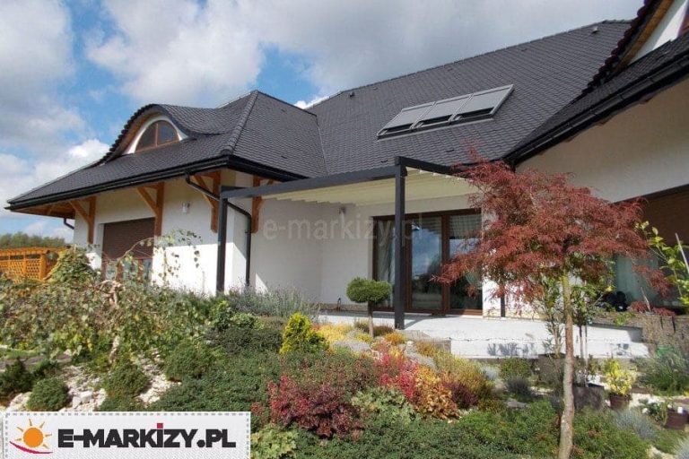 Zdjęcie prezentuje nowoczesną pergolę rzymską Moderno w wersji przyściennej, zamontowaną na tarasie domu jednorodzinnego o białej elewacji i wielospadowym dachu krytym ciemną dachówką. Aluminiowa konstrukcja pergoli w kolorze antracytowym kontrastuje z jasnym, kremowym poszyciem dachu, który jest częściowo zwinięty w kierunku elewacji. System wspiera się na pionowych profilach zamocowanych do betonowej płyty tarasu. Przed tarasem znajduje się bogato zaaranżowany ogród z licznymi krzewami ozdobnymi, bylinami oraz dekoracyjnym drzewkiem o czerwonawych liściach. Dom posiada charakterystyczne detale architektoniczne, takie jak okno typu wole oko oraz okna dachowe z zamontowanymi zewnętrznymi markizami. Całość kompozycji ukazuje integrację nowoczesnego systemu zadaszenia z klasyczną bryłą budynku i otaczającą go roślinnością w słoneczny dzień.