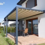 Zdjęcie prezentuje pergolę elektryczną Tarasola Flat o konstrukcji aluminiowej w kolorze antracytowym, zamontowaną do białej elewacji piętrowego budynku. Zadaszenie stanowi jasny, kremowy dach tkaninowy, który jest w pełni rozwinięty, tworząc niemal płaską powierzchnię z delikatnymi, regularnymi liniami profili przeciwwietrznych. Konstrukcja wspiera się na dwóch solidnych, przednich słupach o kwadratowym przekroju, osłaniając taras wyłożony jasnobrązowymi płytkami ceramicznymi imitującymi drewno. W centralnej części tarasu ustawiono drewniany stół z czterema pasującymi krzesłami o konstrukcji metalowej z drewnianym wykończeniem oparć i siedzisk. Duże, przesuwne drzwi tarasowe w brązowych ramach zapewniają bezpośrednie wyjście z wnętrza domu pod zadaszenie. Otoczenie stanowi zadbany trawnik, niskie nasadzenia z traw ozdobnych i lawendy przy krawędzi tarasu oraz rząd tui tworzący żywopłot wzdłuż ogrodzenia. W tle widoczna jest nowoczesna zabudowa jednorodzinna pod czystym, błękitnym niebem.